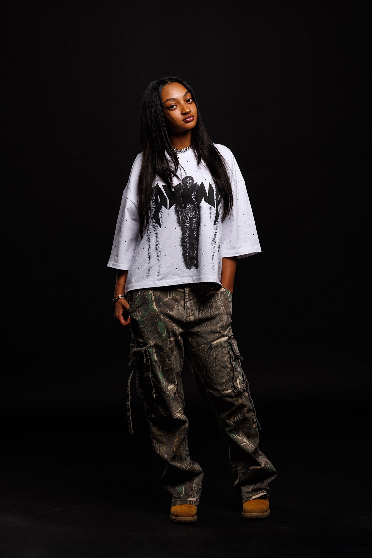 Person wearing a white oversized shirt with black graphics and camouflage pants on a black background