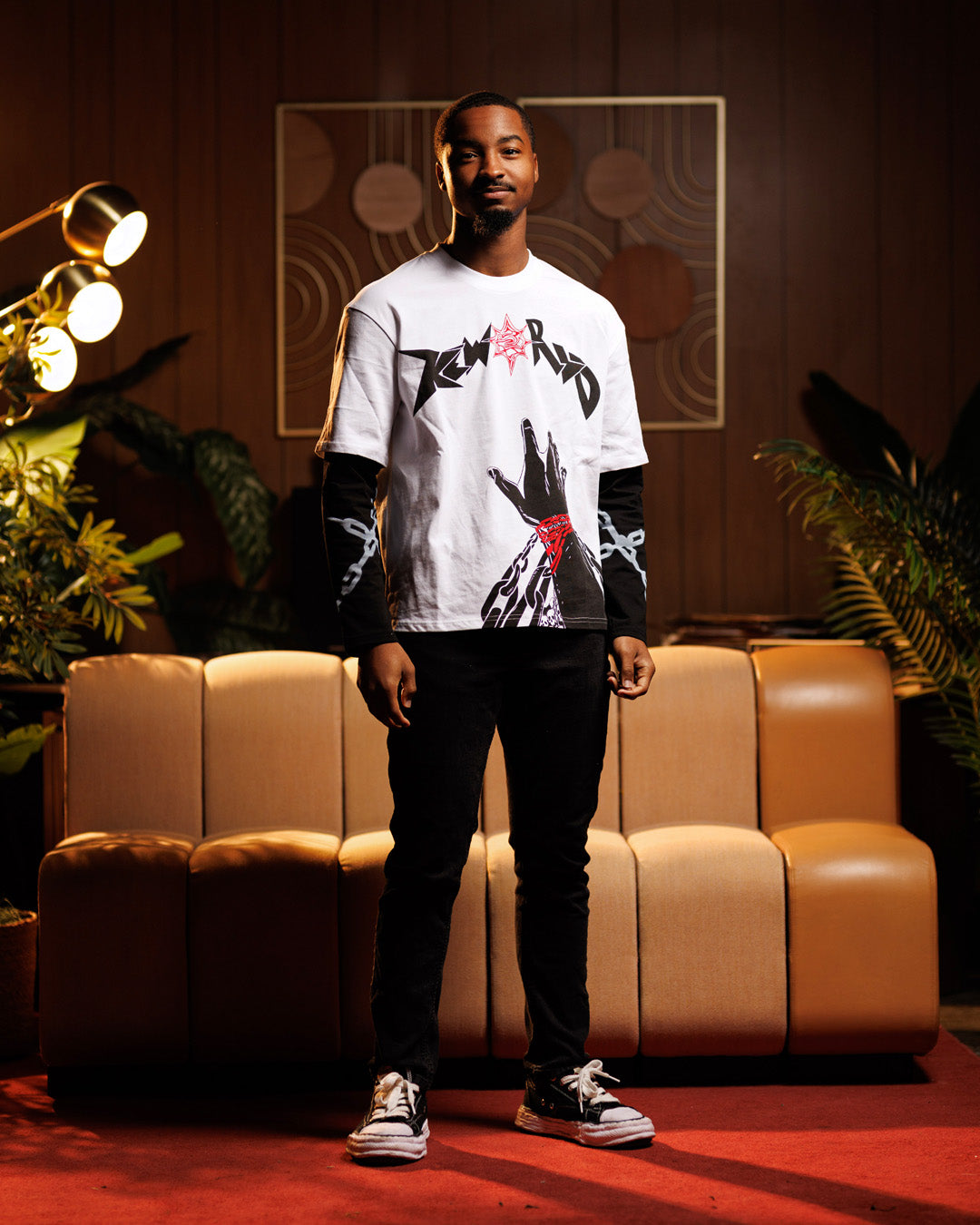 Man wearing a white graphic t-shirt standing in a room with a beige couch and decorative lights.