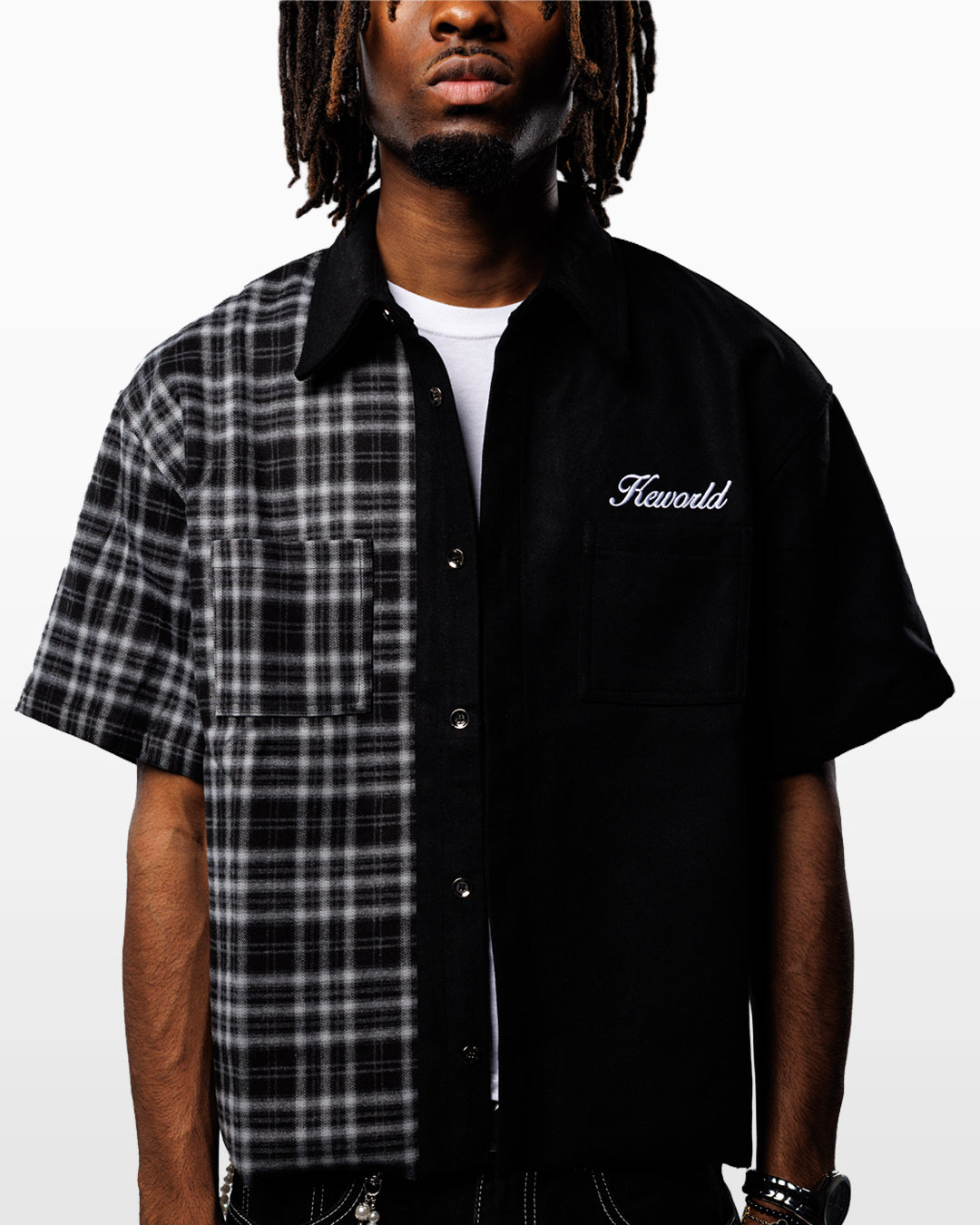 A man wearing a short-sleeve button-up shirt by Keworld, featuring a black and white plaid pattern on the left side and solid black on the right side, with a white t-shirt underneath.