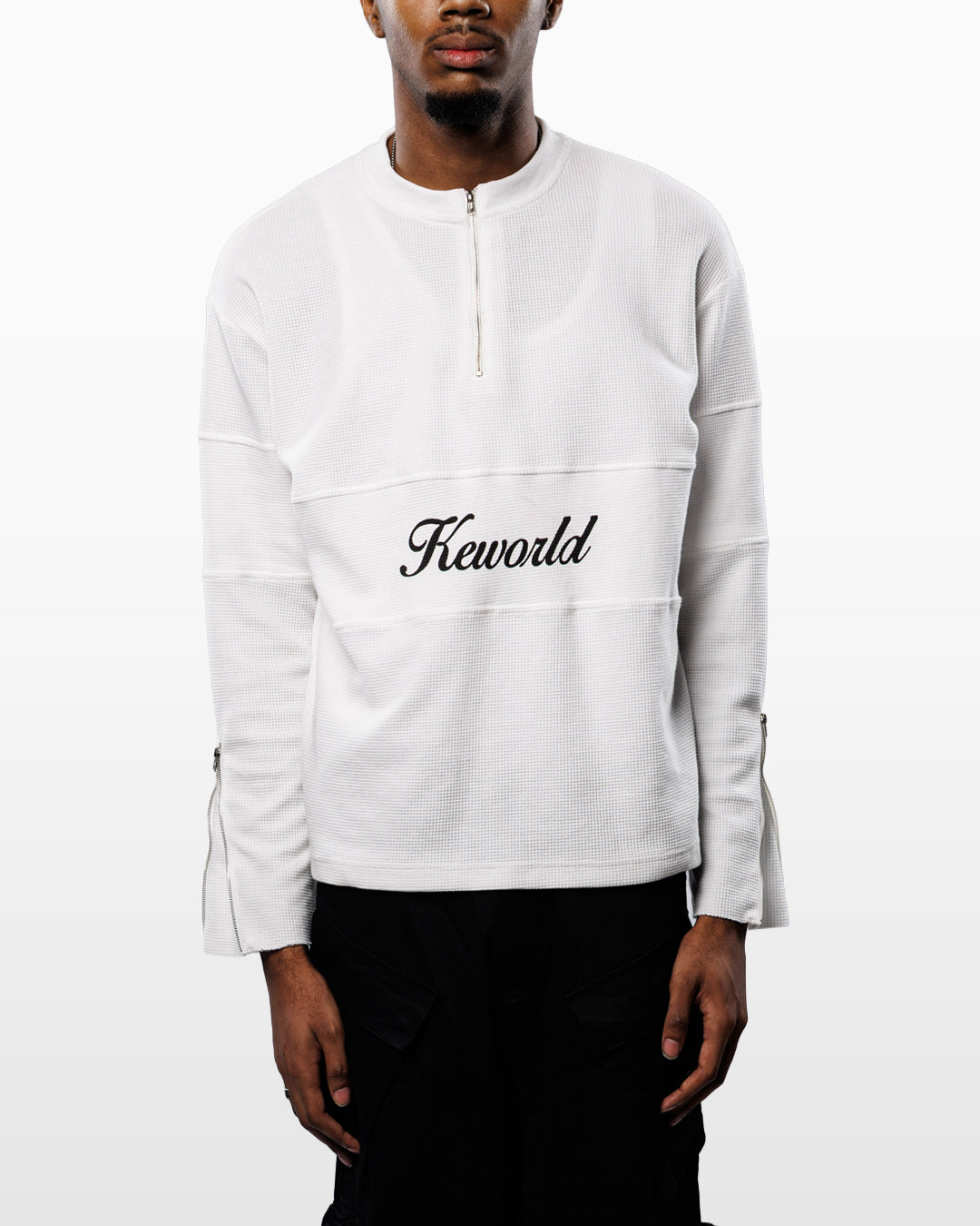 A model wearing a white long-sleeve quarter-zip pullover sweatshirt with 'Kewold' embroidered in black script across the chest, paired with black pants against a light gray background.